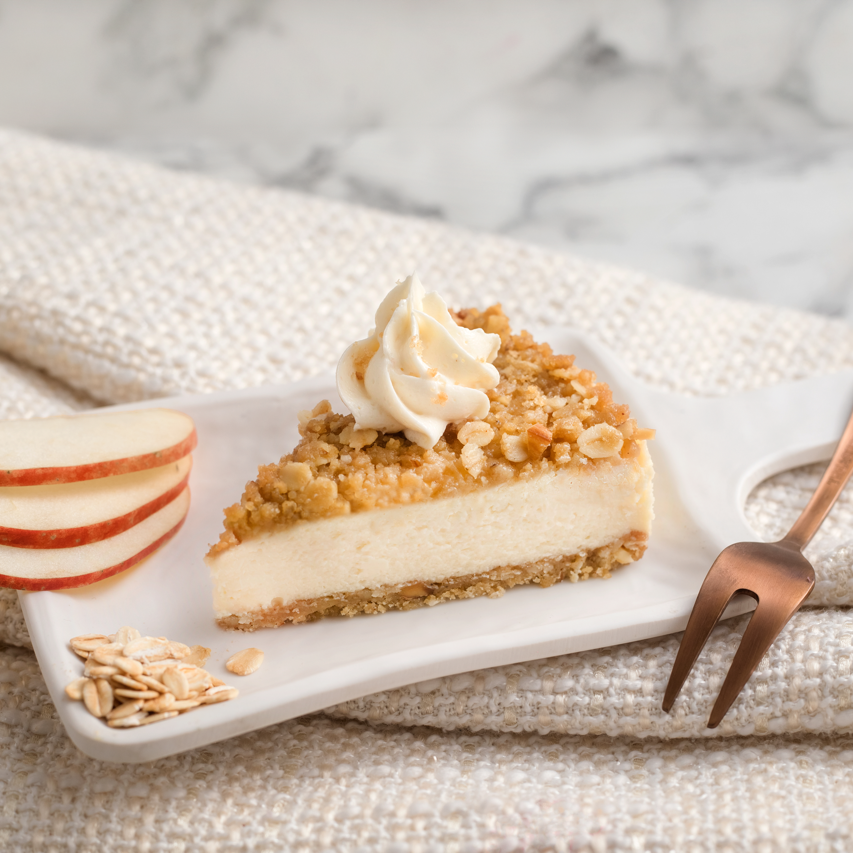 A slice of cheesecake with apple chunks, oats, and almonds, topped with buttercream frosting, on a white plate with a fork and apple slices beside it, placed on a beige napkin.