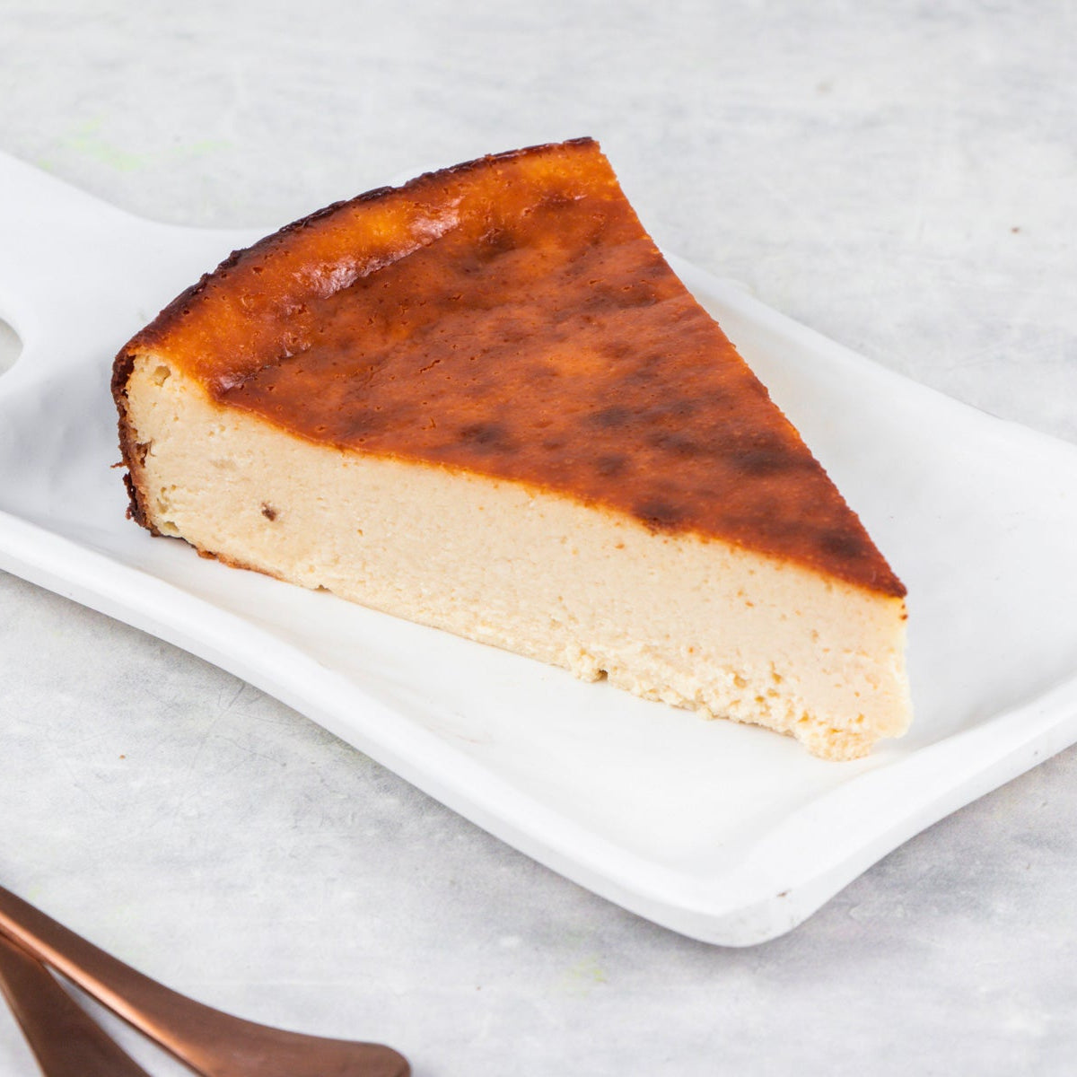 A slice of burnt basquecheesecake on a white plate with a fork and knife to the side, on a marble surface.