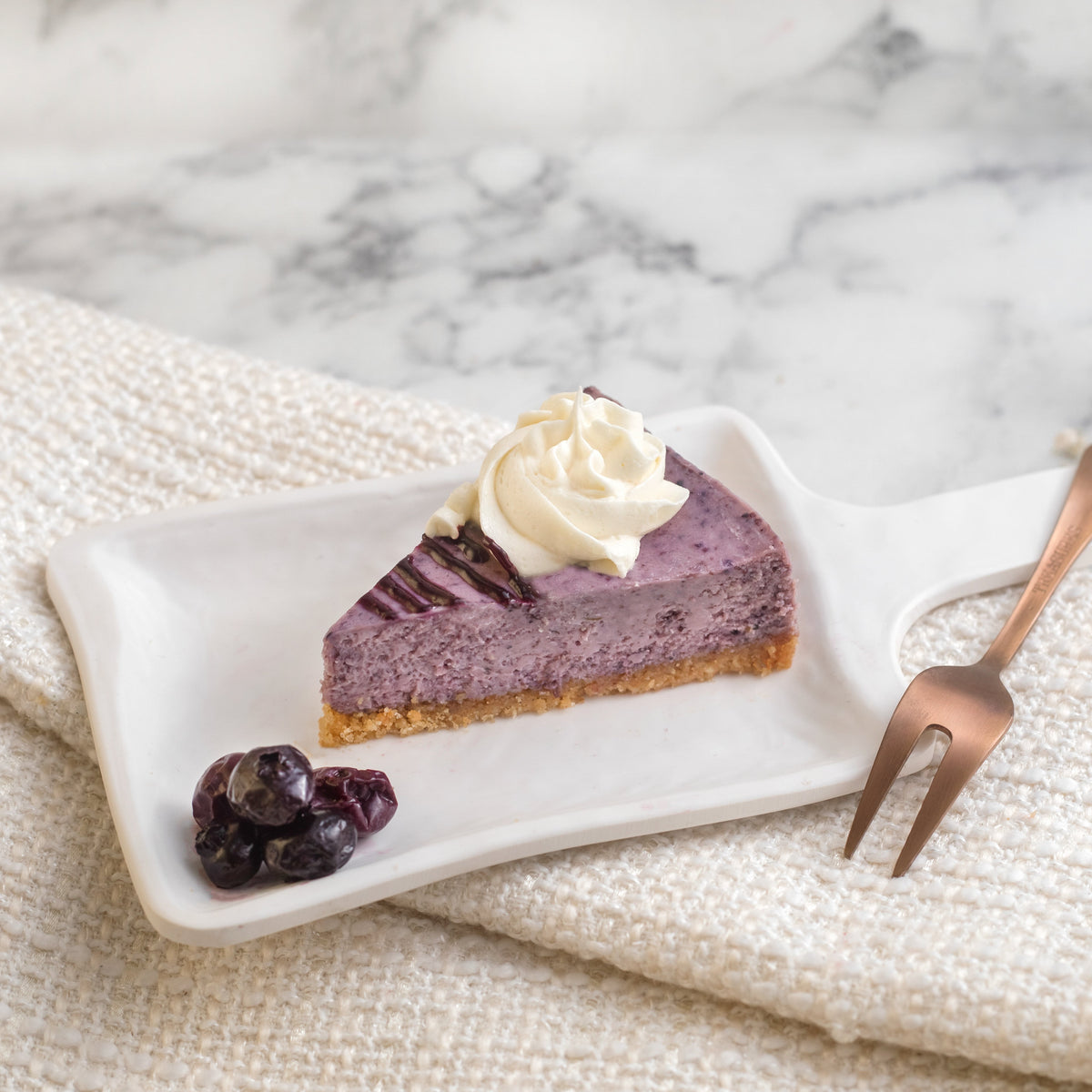 A slice of blueberry cheesecake on a white plate with a dollop of whipped cream on top, a sprinkle of powdered sugar, and a side of fresh blueberries. A fork and a napkin are also visible.