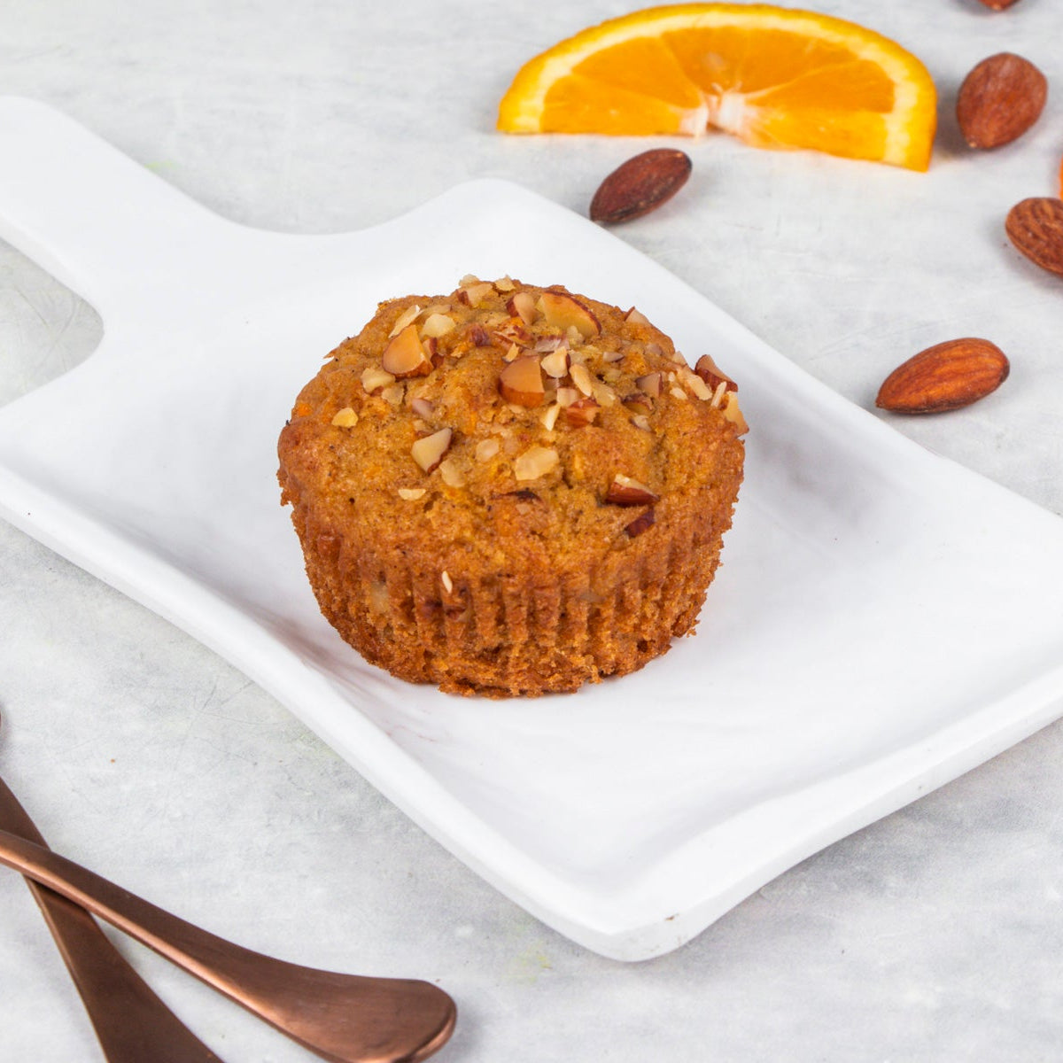 A carrot orange muffin on a white plate with orange zest and nuts scattered around, with a fork and knife on the side.