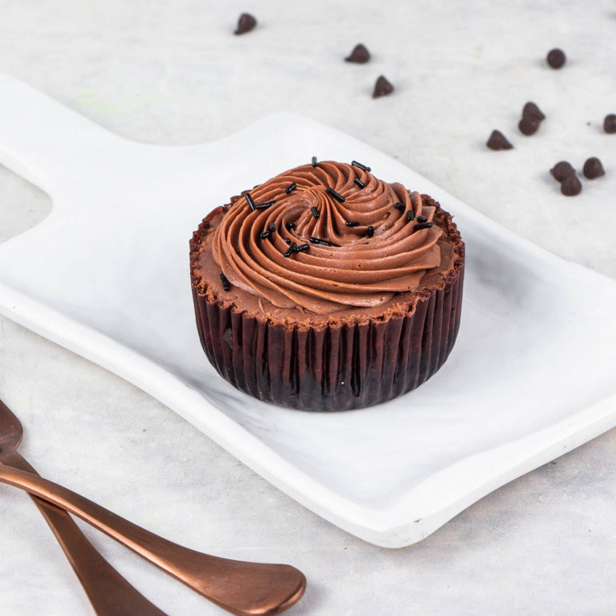A chocolate cheesecake bite with creamy frosting on a white plate with scattered chocolate chips around it and copper-colored utensils in the foreground.