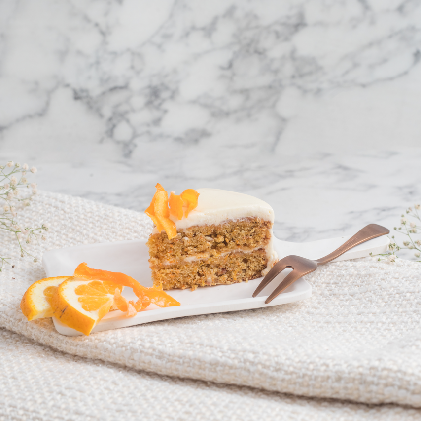 A slice of carrot orange cake with buttercream frosting and decorative orange peel strips on top, placed on a white plate with a fork, against a marble background with a white napkin and decorative flowers.