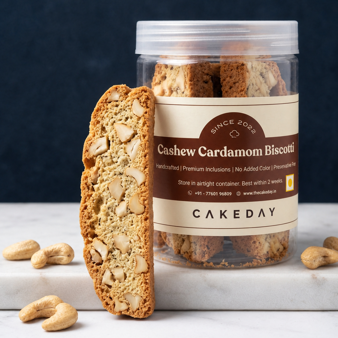 A clear cylindrical jar of Cakeday Cashew Cardamom Biscotti with a beige label, shown on a white marble slab with a single biscotti leaning in front and cashews scattered around. The background is a dark navy blue.