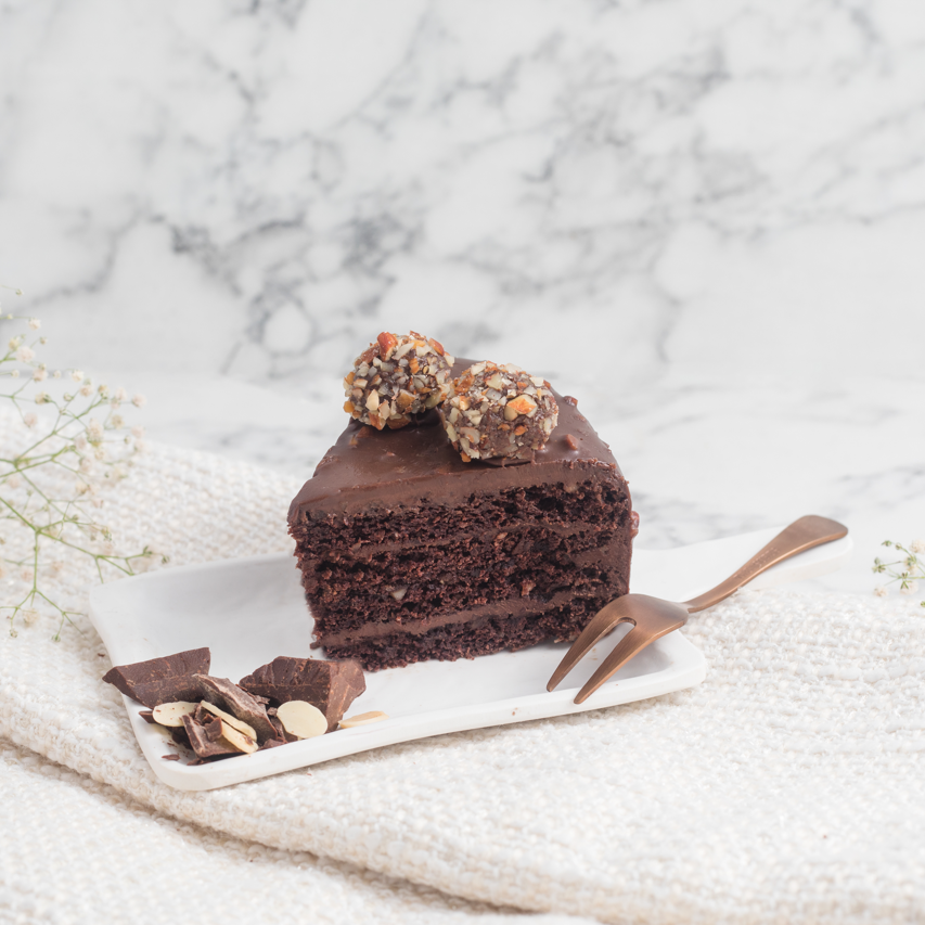 A slice of chocolate almond truffle cake on a plate with a fork, alongside roasted almonds and a white napkin, on a marble surface.