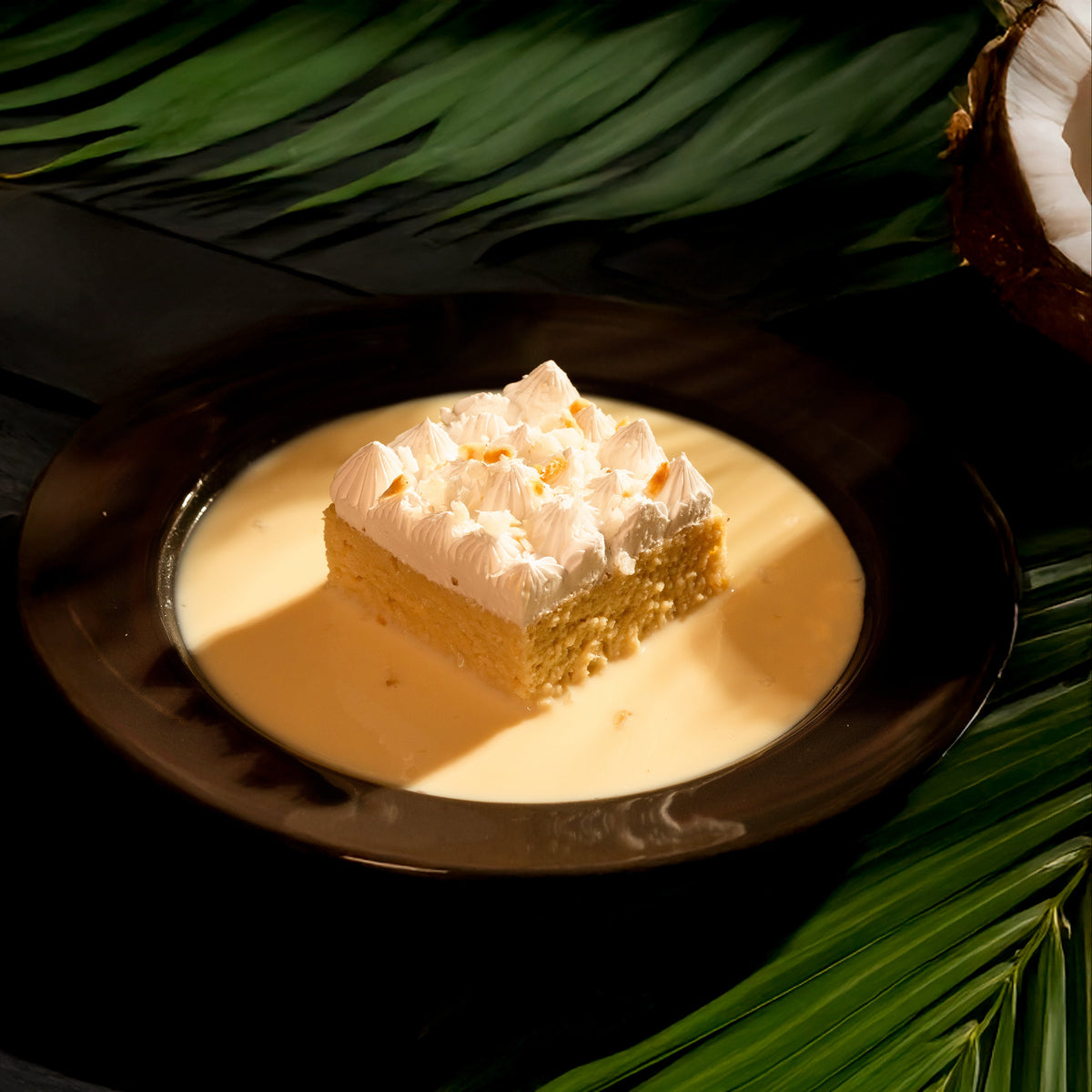 A piece of Coconut Tres Leches cake topped with fresh whipped cream and coconut flakes, served on a plate with a whole cake, coconuts, and leaves in the background.