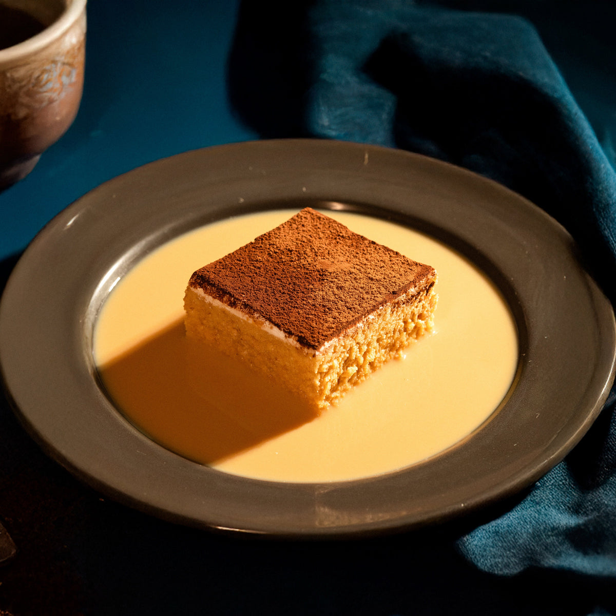 A piece of Coffee Tres Leches cake on a plate with a cup of coffee in the background.