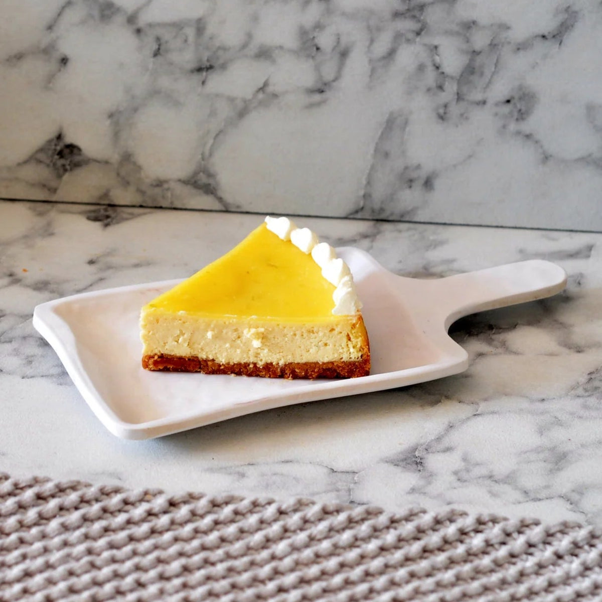 A single slice of lemon cheesecake on a white plate, with a decorative lemon curl on top, placed on a marble surface.