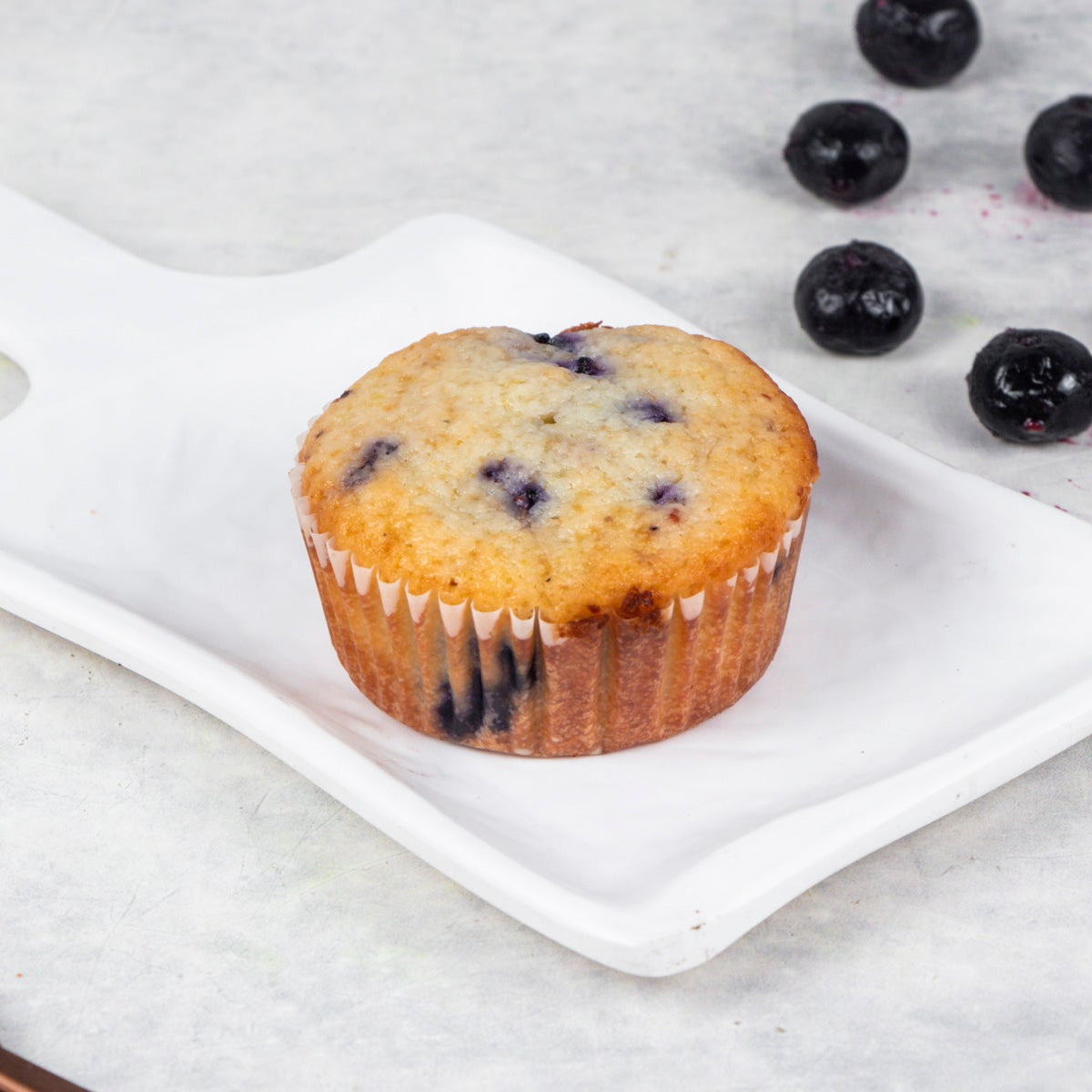 A Lemon Blueberry Muffin on a white plate with blueberries scattered around and a fork to the side, on a marble surface.