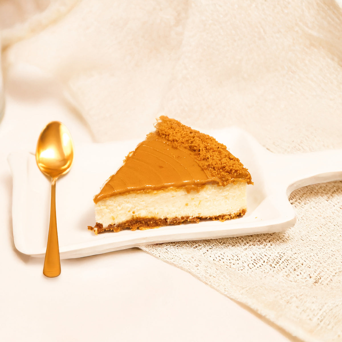 A single slice of Lotus Biscoff Cheesecake on a white plate with a drizzle of caramel on top, accompanied by a gold spoon, on a beige background with a lace tablecloth.
