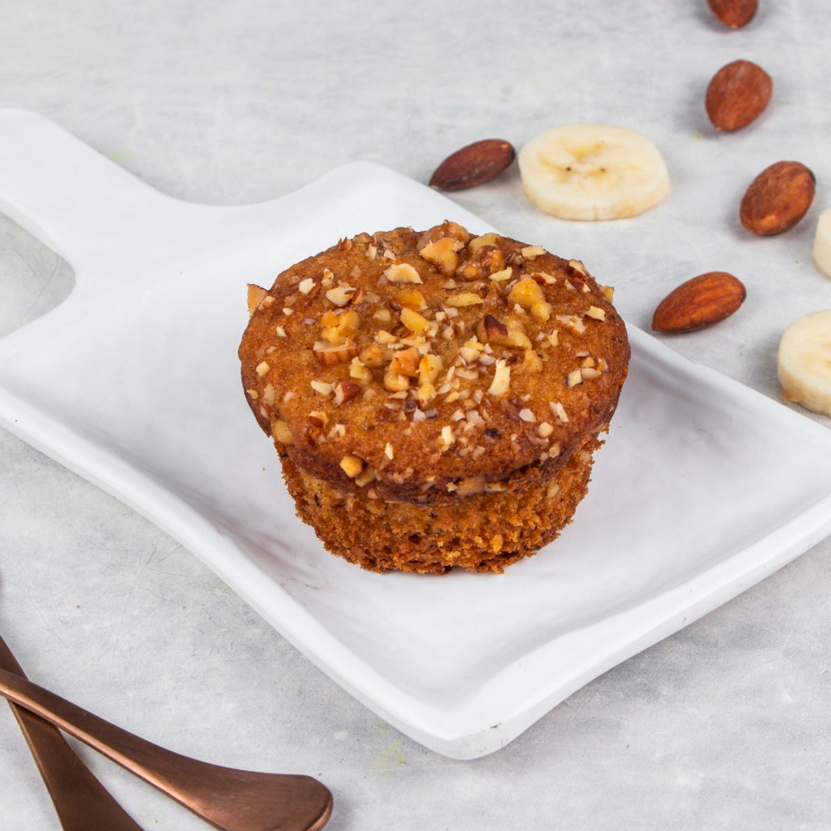 A banana muffin topped with nuts, served on a white plate with bananas and nuts around it, with a grey background.