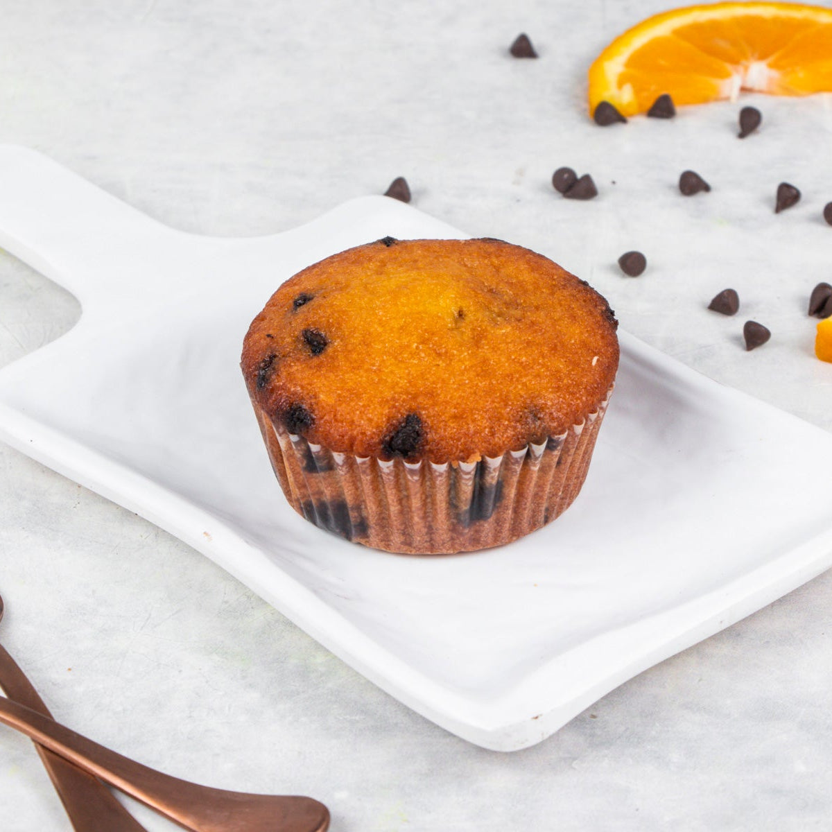 An orange chocolate chip muffin on a white plate with orange zest and chocolate chips scattered around, with a fork and orange slice on the side.