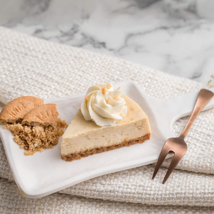 A slice of cheesecake and buttercream frosting, accompanied by a cookie, on a white plate with a fork on the side, presented on a marble surface with a beige cloth.