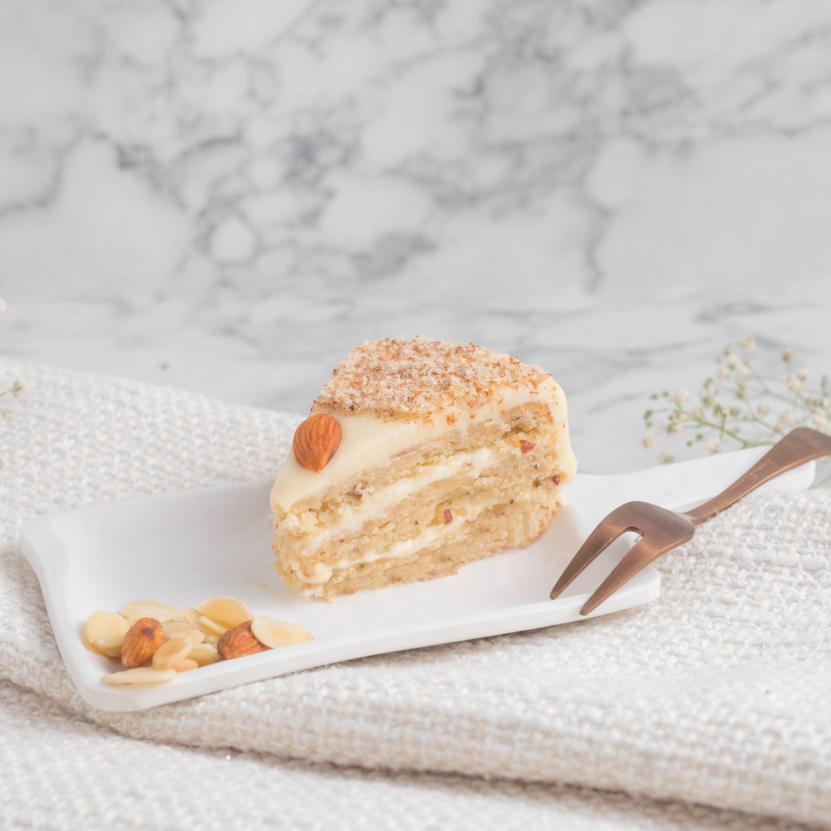 A slice of vanilla almond pastry with roasted almonds and a dusting of powdered sugar on a plate with a fork, against a marble background.