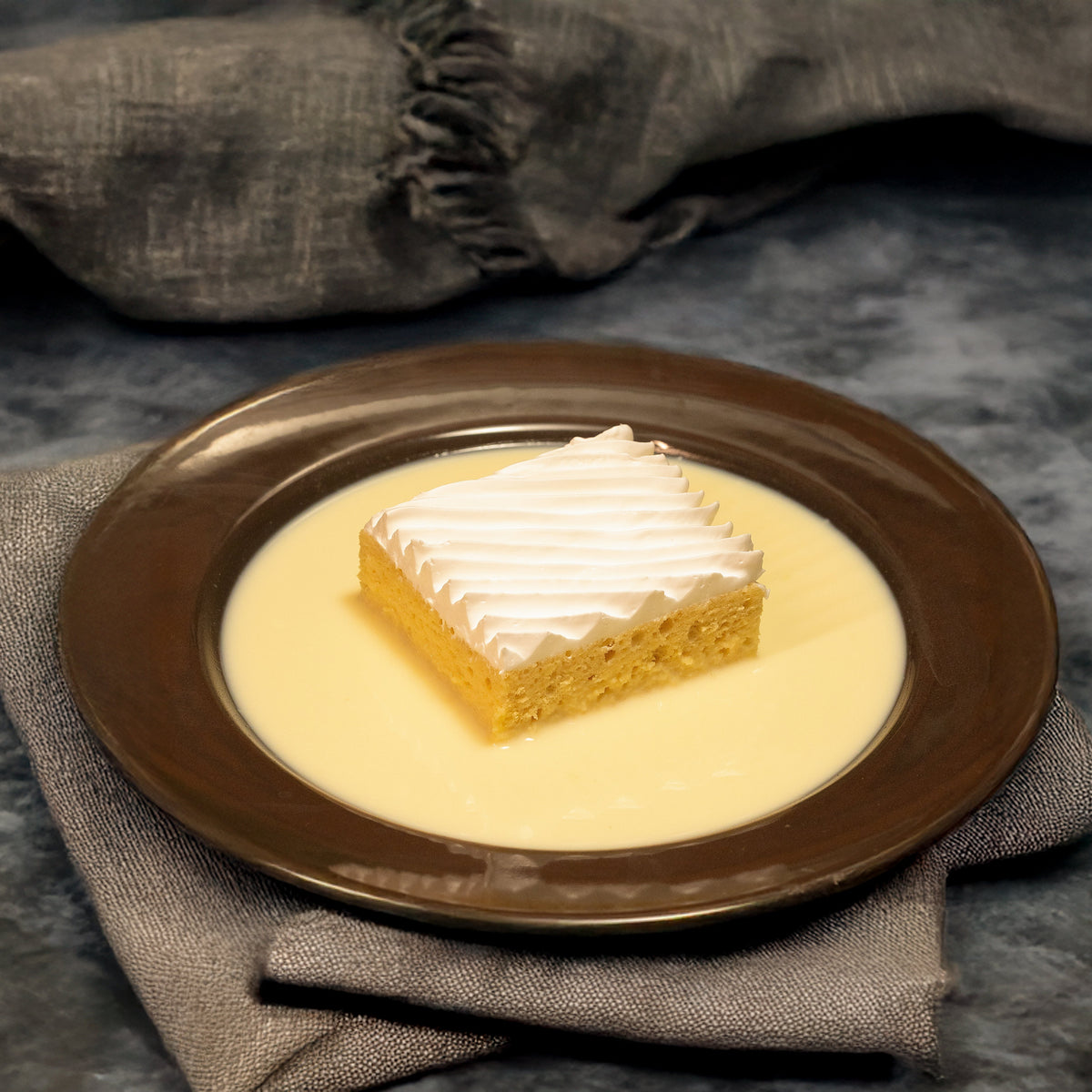 A piece of Tres Leches Cake on a brown plate with a drizzle of white icing on top, accompanied by a jar of milk and a spoon, presented on a grey napkin against a textured background.