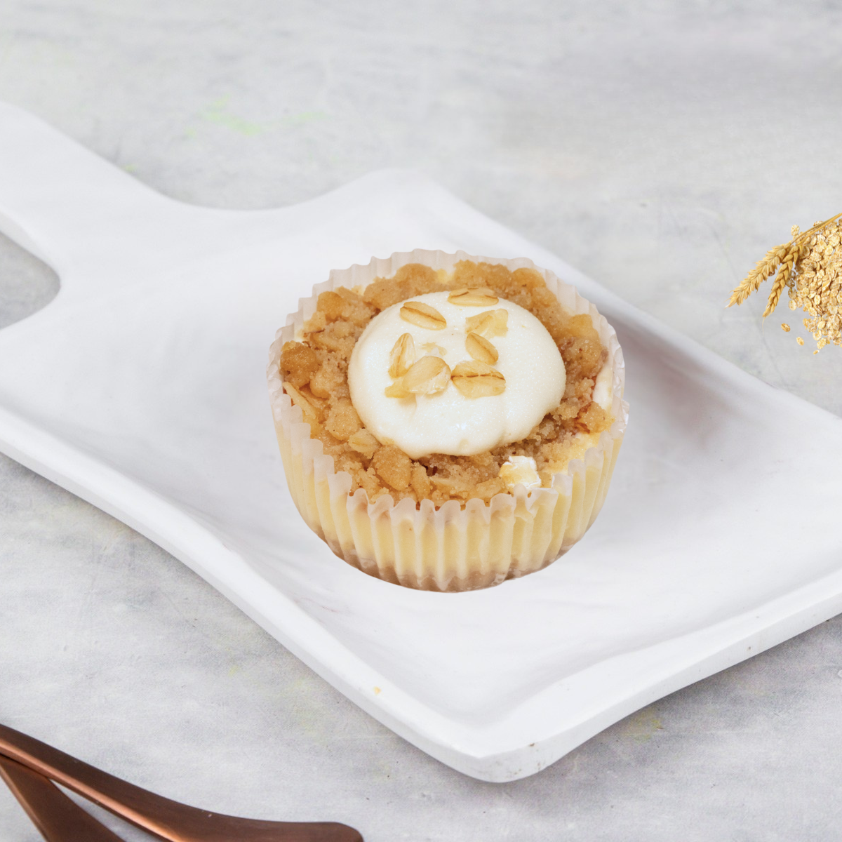 A single cheesecake bite with apple crumble topping and a drizzle of cream on top, served on a white plate with a fork and knife beside it.