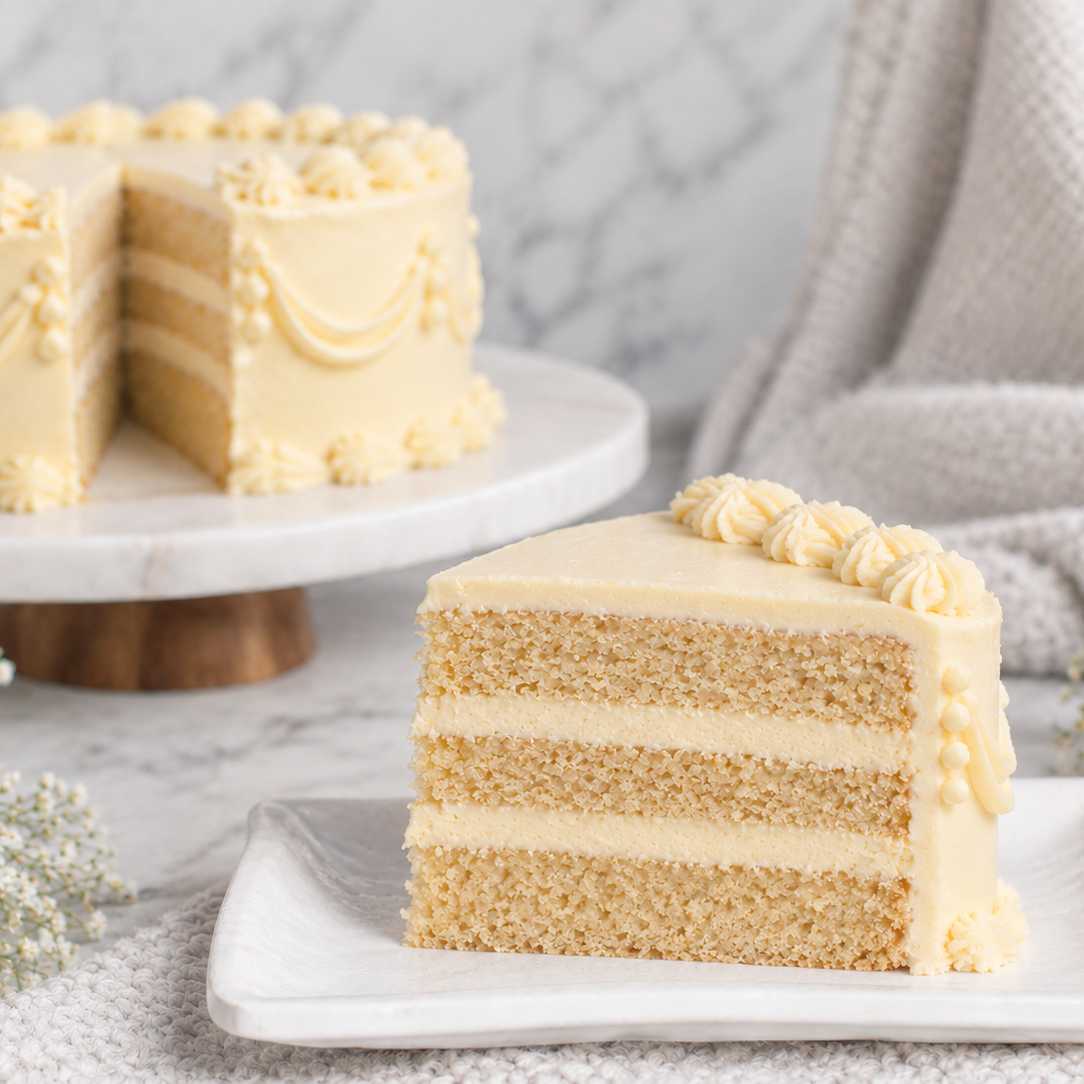 A slice of layered cake with pale yellow frosting sits on a white plate in the foreground, with the full cake on a marble stand blurred in the background. The scene is styled with a soft beige knit cloth and small white flowers on a light surface.