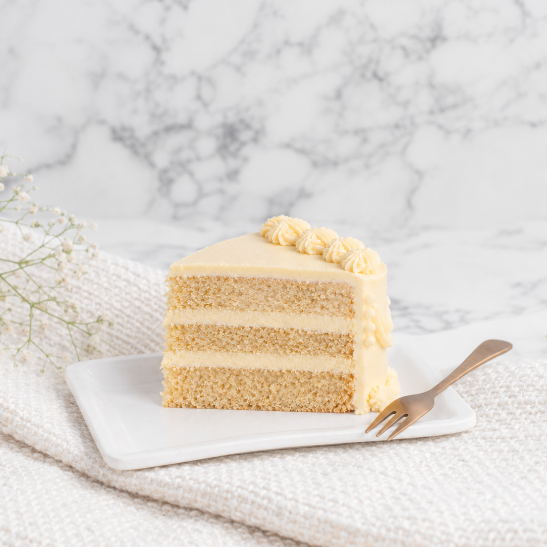 A slice of layered cake with pale yellow frosting sits on a small white plate, with a gold fork beside it on a textured cream fabric against a marble background.