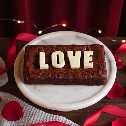 Brownie with 'LOVE' letters on a marble plate surrounded by red ribbons and petals.