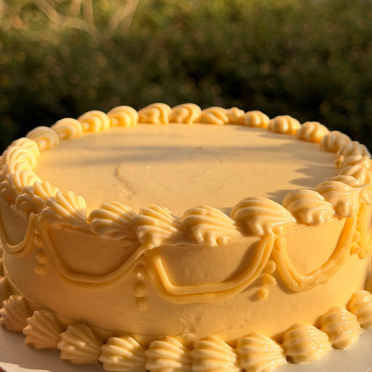 A round yellow cake with piped cream frosting decoration, photographed outdoors with a blurred green garden background.