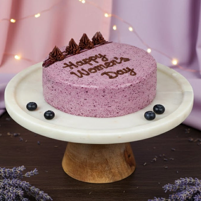 Pink cake with 'Happy Women's Day' on a marble stand against a purple fabric background.
